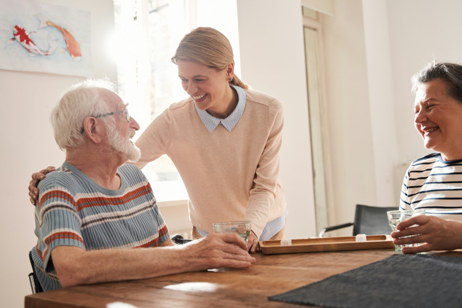 A professional caregiver smiling and assisting an elderly man at a table while a family member looks on, representing respite care for family caregivers in Colorado.