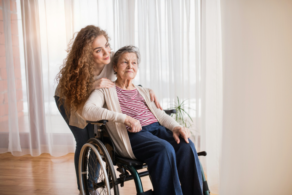 A young woman standing behind an elderly woman in a wheelchair, both looking forward calmly in a bright home setting, representing family caregiving and signs an elderly parent needs help at home.
