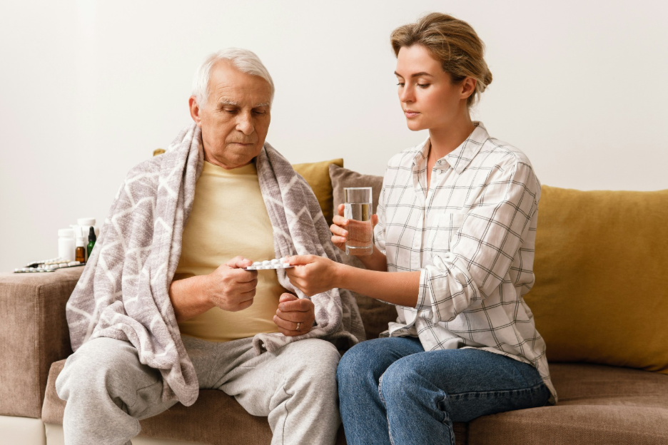 A young woman sitting beside an elderly man on a couch, helping him with medication and a glass of water, representing paid family caregiving in Colorado.